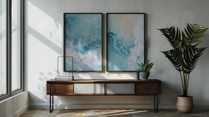 May include: Two framed abstract art prints with blue and white tones hang above a wooden console table. A potted plant and decorative items sit on the table. A large potted plant with green leaves is to the right of the table.