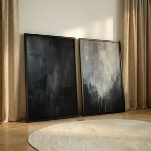May include: Two large abstract paintings in black and white, leaning against a wall. The artwork features textured brushstrokes and a dark, moody aesthetic. The paintings are framed in black and rest on a wooden floor, with a cream-colored rug in the foreground.