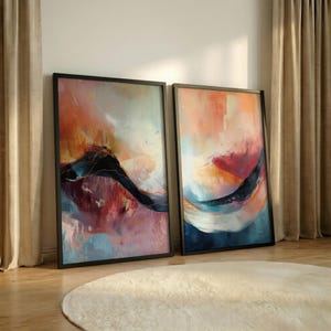May include: Two framed abstract art prints. The artwork features a blend of colors, including orange, red, blue, and black, with a flowing, dynamic composition. The frames are black, and the art is displayed on a wooden floor.