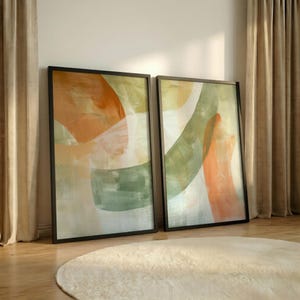 May include: Two framed abstract art prints. Each print features a blend of orange, green, and white brushstrokes on a light background, encased in a black frame. The artwork leans against a wall, with a cream-colored rug in the foreground.