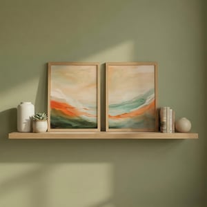 May include: Two framed abstract landscape paintings with orange, green, and beige tones. The artwork is displayed on a light wood shelf, with a white vase, a small succulent in a pot, books, and a small spherical vase.