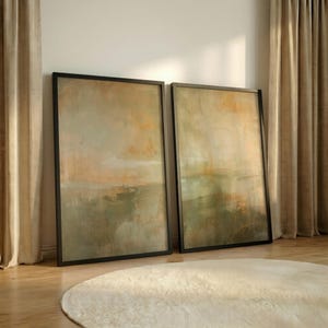 May include: Two rectangular framed wall art pieces featuring abstract brushwork in muted tones of sage green, soft orange, and beige. The paintings display vertical paint drips and blended color gradients, each enclosed in a thin black frame.
