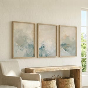 May include: Three framed abstract art prints. Each print features a soft, blended design with shades of blue, cream, and beige. The artwork is encased in light-colored wooden frames, creating a cohesive set for wall decor.