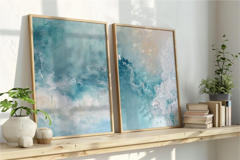 May include: Two framed abstract art prints with ocean-inspired designs in shades of blue, white, and beige. The artwork is displayed on a wooden shelf with decorative plants and books. The frames are made of light-colored wood.