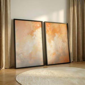 May include: Two framed abstract art prints. The artwork features a blend of orange, peach, and white hues, creating a soft, cloud-like effect. The frames are black, and the prints are leaning against a wall. The room has a neutral color palette.