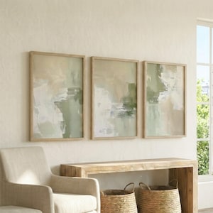 May include: Three framed abstract art prints in shades of green, beige, and white. The artworks are displayed on a light-coloured wall. The frames are light wood, above a wooden console table. The overall aesthetic is neutral.