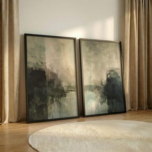May include: Two framed abstract art prints. The artwork features a blend of dark blues, greens, and neutral tones, with visible brushstrokes and a textured appearance. The frames are black, and the art leans against a wall near a beige curtain and a round, cream-colored rug.