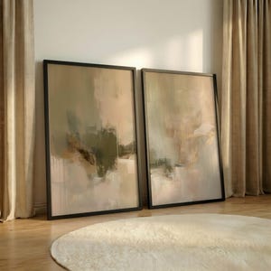 Könnte beinhalten: Zwei gerahmte abstrakte Kunstdrucke. Das Kunstwerk zeigt eine Mischung aus neutralen Farben, darunter Beige, Olivgrün und Gold, mit sichtbaren Pinselstrichen und einem strukturierten Aussehen. Die Rahmen sind schwarz, und die Kunst lehnt an einer Wand. Ein cremefarbener Teppich ist im Vordergrund.