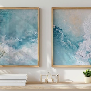 May include: Two framed abstract art prints with a light wood frame. The artwork features shades of blue and white, resembling ocean waves. The prints are displayed above a wooden shelf with plants and books.