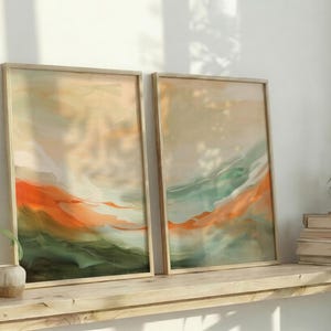 May include: Two framed abstract art prints. The artwork features a blend of orange, green, and beige hues, creating a landscape-like composition. The frames are light-coloured wood, and the prints are displayed on a wooden shelf with decorative items.