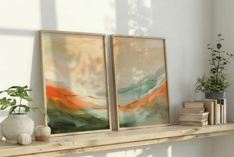 May include: Two framed abstract art prints. The artwork features a blend of orange, green, and beige hues, creating a landscape-like composition. The frames are light-coloured wood, and the prints are displayed on a wooden shelf with decorative items.