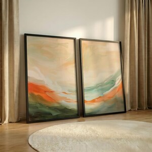 May include: Two framed abstract art prints. Each piece features a blend of orange, green, and beige hues, creating a landscape-like composition. The artwork is encased in a black frame, leaning against a wall.