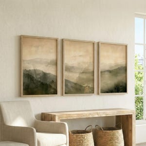 Peut inclure: Trois impressions d'art encadrées représentant un paysage de montagne brumeux dans des tons de vert, de gris et de beige. L'œuvre est exposée sur un mur blanc au-dessus d'une console en bois. Les cadres sont de couleur marron clair.