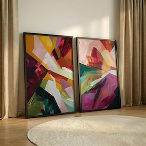 May include: Two framed abstract art prints. The artwork features a vibrant mix of colors, including yellow, orange, red, green, and purple, in a geometric, layered design. The frames are black, and the prints are leaning against a wall.