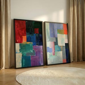 Puede incluir: Dos pinturas abstractas enmarcadas con marcos negros. Las pinturas presentan una mezcla de colores, incluyendo rojo, verde, azul, morado y blanco. La obra de arte está apoyada contra una pared cerca de una cortina beige.