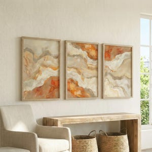 May include: Three framed abstract art prints. Each print features a flowing design with shades of orange, beige, and gray, resembling a landscape. The frames are made of light-colored wood. The artwork is displayed on a white wall.