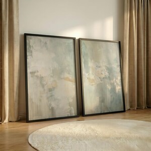 May include: Two framed abstract art prints with a muted color palette of blues, greens, and creams. The artwork is displayed against a neutral wall, with a beige rug and curtains in the background. The frames are black.