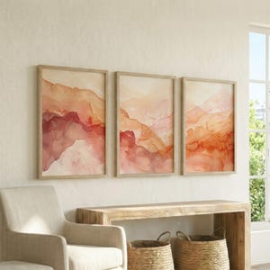 May include: A set of three rectangular wall art prints featuring abstract watercolour landscapes in shades of terracotta, peach, and cream. Each print is encased in a light-toned wooden frame, displaying fluid, layered mountain-like shapes.