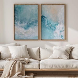 May include: Two framed abstract art prints with blue and white tones hang above a cream-colored sofa with several pillows and a tan throw blanket. A wooden side table holds a vase of dried flowers.
