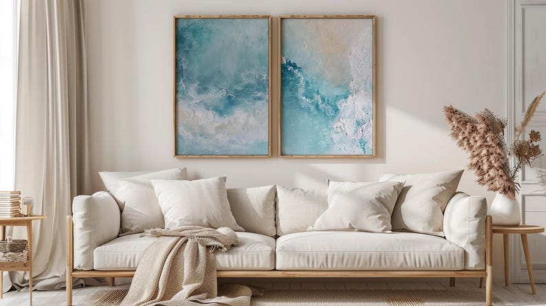 May include: Two framed abstract art prints with blue and white tones hang above a cream-colored sofa with several pillows and a tan throw blanket. A wooden side table holds a vase of dried flowers.