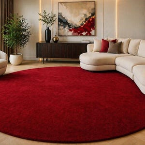 May include: A large, circular, solid deep red area rug with a textured surface. The rug is positioned on a light-colored wooden floor in a room featuring a cream-colored sectional sofa, a matching armchair, and dark wood furniture pieces.