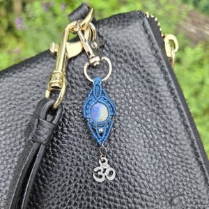 May include: A blue macrame keychain with a round, iridescent stone and a silver-colored Om charm. The keychain is attached to a black leather purse with gold-colored hardware. The background is blurred green foliage.