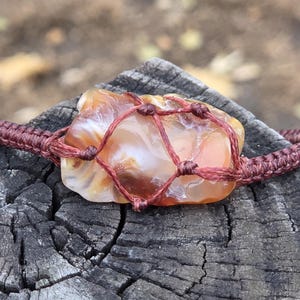May include: A natural stone pendant, featuring a polished, orange and white agate stone, is secured within a woven, dark red macrame cord. The stone is approximately 5 cm long. The cord forms a bracelet, showcasing a rustic, earthy aesthetic.