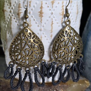 May include: A pair of dangle earrings featuring an antique brass-toned teardrop-shaped metal component with an intricate filigree cutout pattern. Attached to the base of each metal piece are several dark gray thread loops in a fringe design.