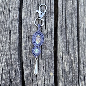 May include: A handcrafted keychain featuring a macrame design in a dark blue color. The keychain has two oval-shaped abalone shell accents and a silver-colored teardrop charm. A silver-colored clasp is attached at the top.