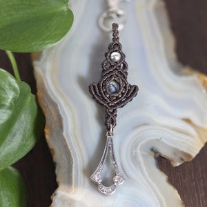 Op de afbeelding: Een donkerbruine macramé sleutelhanger met een centrale iriserende cabochon en een kleine zilveren kraal. Het macramé gedeelte is aan de bovenkant bevestigd aan een zilverkleurige metalen karabijnhaak en aan de onderkant aan een zilverkleurige hanger met heldere steentjes.