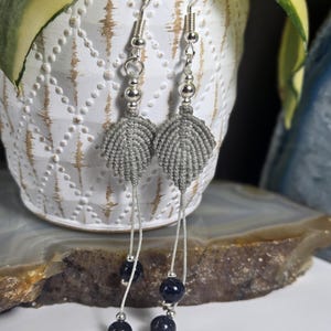 May include: A pair of dangle earrings featuring grey macrame leaf-shaped charms suspended from silver-toned hooks. Each earring includes a small silver bead above the leaf and two dark blue, glittery round beads hanging on thin cords below.