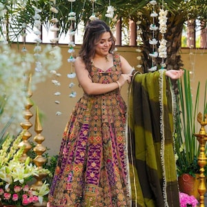 Puede incluir: Vestido sin mangas estampado en tonos verde oliva, morado y dorado. El vestido tiene una falda amplia y se combina con un chal verde. La imagen está tomada al aire libre con decoraciones florales.