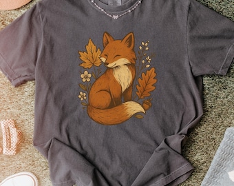 May include: A dark gray t-shirt featuring a cartoon fox illustration. The fox is brown and white, surrounded by autumn leaves and flowers. The shirt has a crew neck and short sleeves. The shirt is made of a soft material.