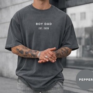 May include: A dark gray, oversized t-shirt with the text "BOY DAD EST. 2026" printed in white. The shirt has a crew neck and short sleeves. The model is wearing gray and white plaid pants.