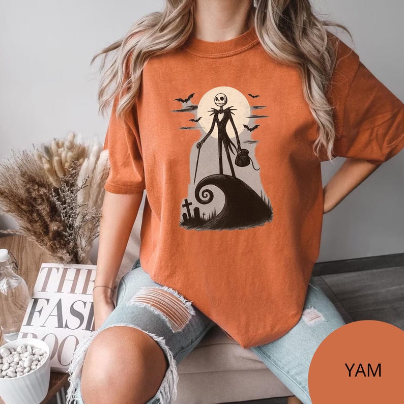 May include: Orange t-shirt with a graphic of Jack Skellington from The Nightmare Before Christmas. The design includes a full moon, bats, and a swirling hill with a tombstone. The shirt is a solid orange colour with the colour name "YAM" at the bottom.
