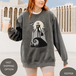 May include: Dark grey sweatshirt with a graphic of Jack Skellington from The Nightmare Before Christmas. The design includes a full moon, bats, and a swirling hill. The sweatshirt has a crew neck and long sleeves.