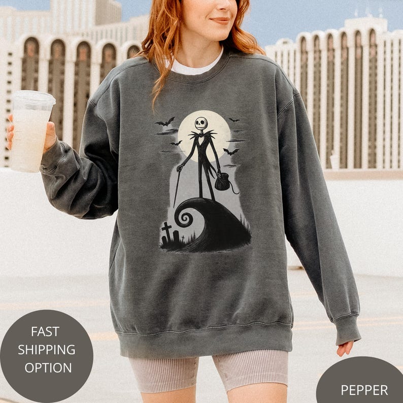 May include: Dark grey sweatshirt with a graphic of Jack Skellington from The Nightmare Before Christmas. The design includes a full moon, bats, and a swirling hill. The sweatshirt has a crew neck and long sleeves.
