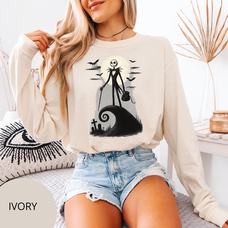 May include: Ivory long-sleeved sweatshirt with a black and white illustration of Jack Skellington from The Nightmare Before Christmas. The design shows Jack on a hill with a full moon, bats, and a tombstone. The word "IVORY" is printed at the bottom.