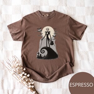 May include: Espresso-coloured t-shirt featuring a graphic of Jack Skellington from The Nightmare Before Christmas. The design shows Jack standing on a hill with a full moon, bats, and a tombstone. The shirt is a crew neck and has short sleeves.
