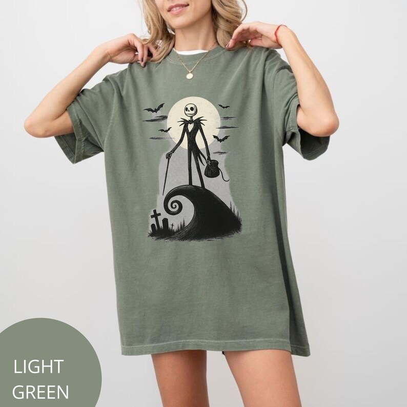 May include: Light green t-shirt with a graphic of Jack Skellington from The Nightmare Before Christmas. The design includes a full moon, bats, and a swirling hill. The shirt has a relaxed fit.