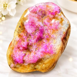 Puede incluir: Un espécimen mineral pulido que presenta un centro cristalino de color magenta vibrante rodeado por una corteza exterior lisa de color tostado. La piedra tiene una forma irregular y redondeada con una textura brillante y motas oscuras.