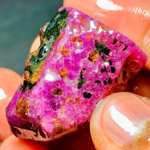 Pode incluir: Uma pedra preciosa bruta e não polida com uma cor base magenta e rosa vibrante. A pedra apresenta superfícies irregulares e texturizadas com inclusões minerais verde-escuras e bronze metálico incrustadas na sua estrutura cristalina.