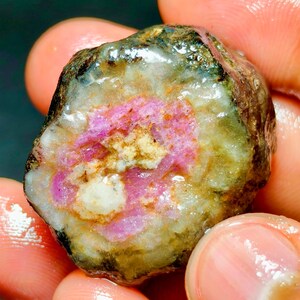 Puede incluir: Una piedra preciosa en bruto y sin tallar con un núcleo rosa vibrante rodeado de capas minerales de color blanco translúcido y marrón terroso. La piedra tiene una textura superficial húmeda y brillante con una forma irregular y redondeada.