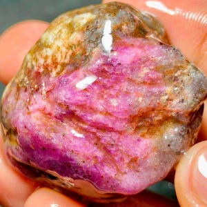 Pode incluir: Uma pedra preciosa bruta e polida com tons vibrantes de magenta e rosa misturados com inclusões minerais castanhas e cinzentas. A pedra tem uma textura de superfície húmida e brilhante com estruturas cristalinas naturais visíveis.