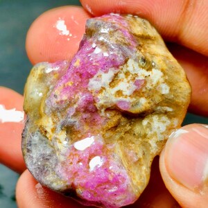 Puede incluir: Una piedra preciosa en bruto y sin pulir que presenta una mezcla de secciones cristalinas de color rosa magenta vibrante incrustadas en una matriz texturizada de color beige terroso y blanco. La superficie de la piedra parece húmeda y brillante, resaltando el contraste entre los depósitos minerales de color rosa brillante y el material rocoso opaco y desgastado que la rodea.