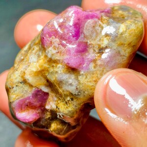 Puede incluir: Una piedra preciosa en bruto y sin pulir que presenta inclusiones cristalinas de color magenta y rosa vibrante incrustadas en una matriz terrosa y texturizada de depósitos minerales de color canela, blanco y translúcido, con un acabado brillante.