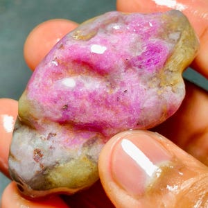 May include: A raw, unpolished gemstone featuring a vibrant magenta and pink center with translucent, pale yellow and grey edges. The stone has a wet, glossy surface texture with visible crystalline inclusions and natural, irregular contours.