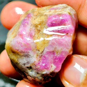 Puede incluir: Una piedra preciosa en bruto y sin pulir que presenta secciones cristalinas de color magenta y rosa vibrante incrustadas en una matriz mineral texturizada de color tostado y gris. La superficie de la piedra parece húmeda y brillante, resaltando el contraste entre la roca opaca y las formaciones minerales rosas, translúcidas y dentadas.