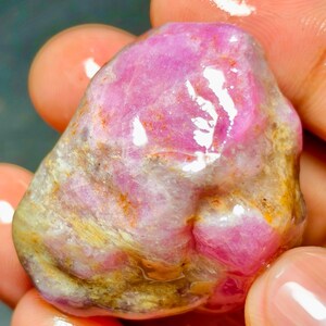 以下が含まれることがあります： 鮮やかなマゼンタとピンクのカラーパレットが特徴の、未加工で磨かれたルビーの宝石。天然のインクルージョン、土のような茶色の鉱物堆積物があり、光沢のある濡れたような表面の質感が、不規則で丸みを帯びた形状全体に光を反射させています。