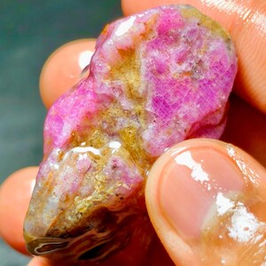 Puede incluir: Una piedra preciosa en bruto y sin pulir con una estructura cristalina de color magenta y rosa vibrante, que presenta inclusiones minerales de color marrón dorado natural y una textura superficial húmeda y brillante que resalta su forma orgánica.